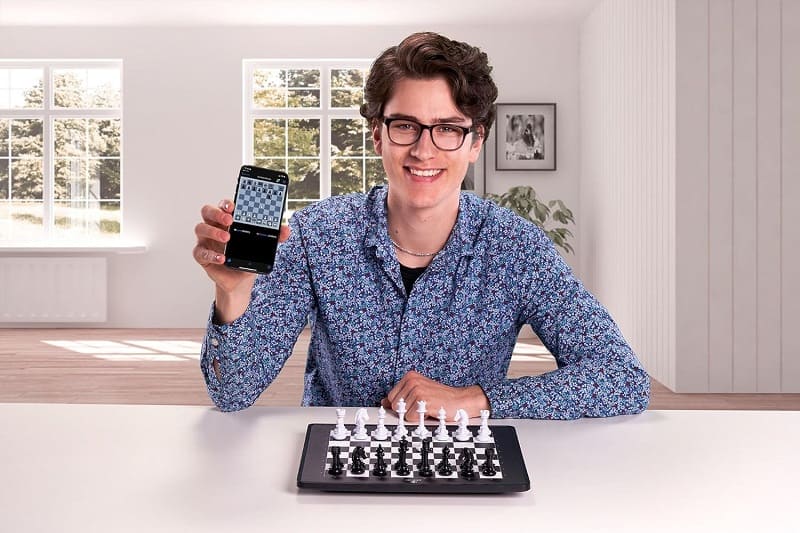 Giovane sorridente mostra telefono con app scacchi, in una stanza luminosa.
